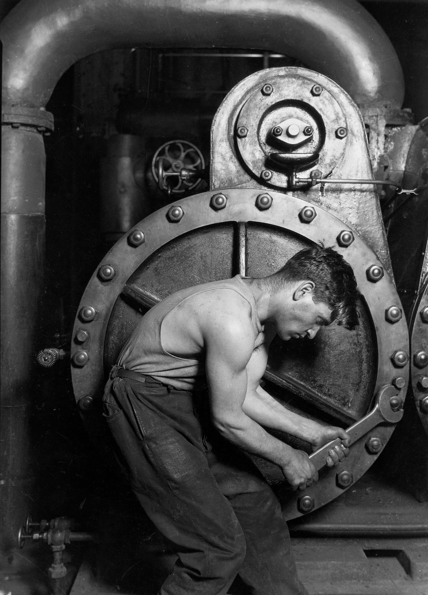 A mechanic bends over a large steam pump with a wrench in Lewis Hine's 1920 photograph